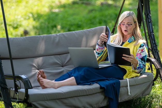 Senior woman with laptop and documents working in garden on rocking couch green home office concept