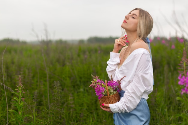 Sensuelle belle fille dans une chemise blanche et avec des ongles brillants roses se tient dans le champ tenant un panier avec des fleurs violettes.