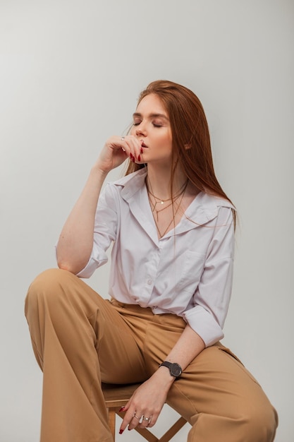 Sensuelle douce belle fille rousse aux longs cheveux roux dans des vêtements élégants avec une chemise blanche et un pantalon est assis sur une chaise en bois