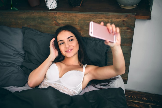 Sensuelle jeune femme en sous-vêtements blancs sur lingerie noire faisant selfie à l'aide d'un smartphone et souriant en position couchée sur le lit.