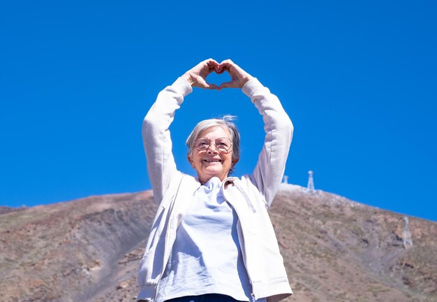 Photo sentiment de liberté pour une femme senior souriante aux cheveux blancs debout sur fond de montagne en forme de coeur avec ses mains