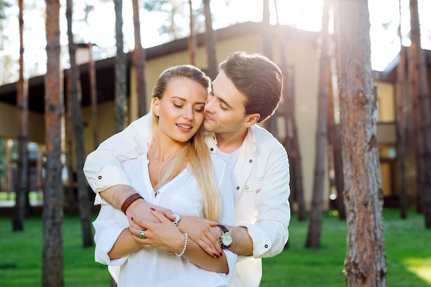 Des sentiments romantiques. Positif beau jeune homme serrant sa femme bien-aimée tout en se tenant avec elle dans les bois