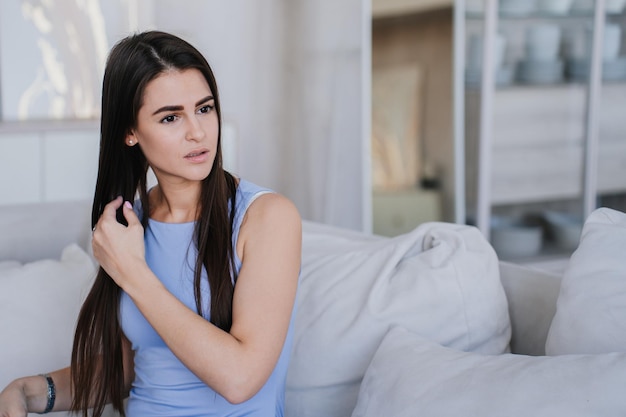 Sérieuse belle femme brune en chemisier lilas assise sur un canapé avec des coussins contre le salon