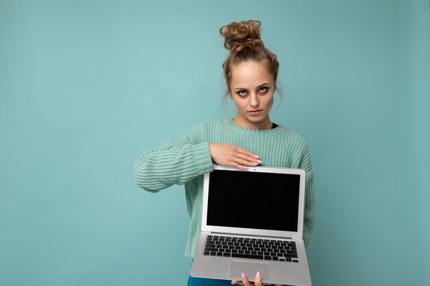 Sérieuse belle jeune femme blonde foncée aux cheveux bouclés rassemblés en regardant la caméra tenant l'ordinateur