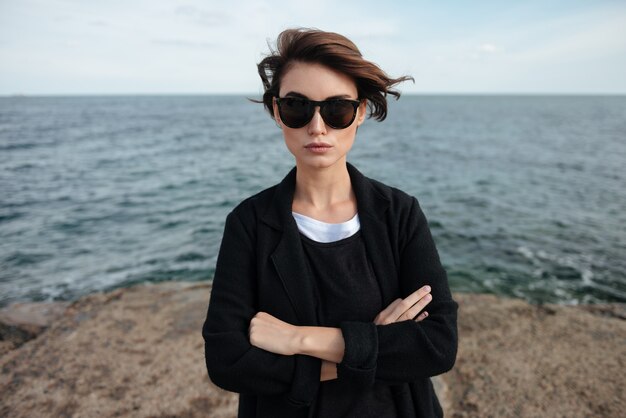 Sérieuse belle jeune femme à lunettes de soleil debout avec les bras croisés au bord de la mer