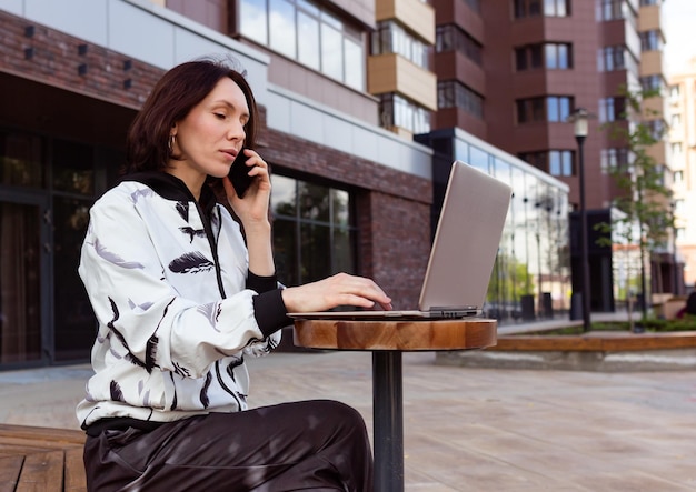 Sérieuse belle jeune femme tapant sur un ordinateur portable et parlant au téléphone dans la rue de la ville moderne