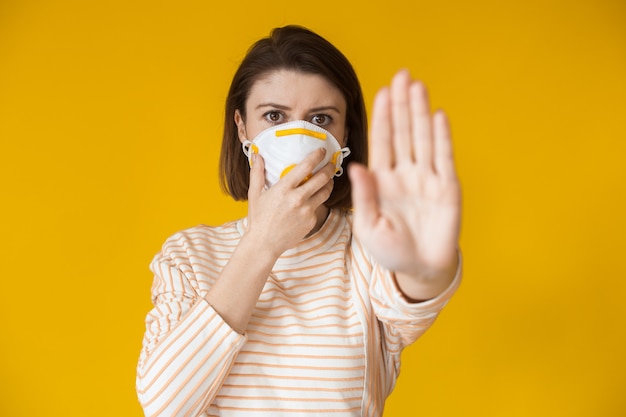 Sérieuse dame de race blanche montrant le panneau d'arrêt tout en portant un masque avec filtre sur un mur jaune