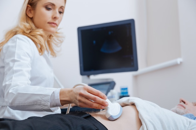 Photo sérieuse femme médecin attrayante travaillant dans la clinique tout en assurant une surveillance par ultrasons de l'estomac et en utilisant un appareil à ultrasons