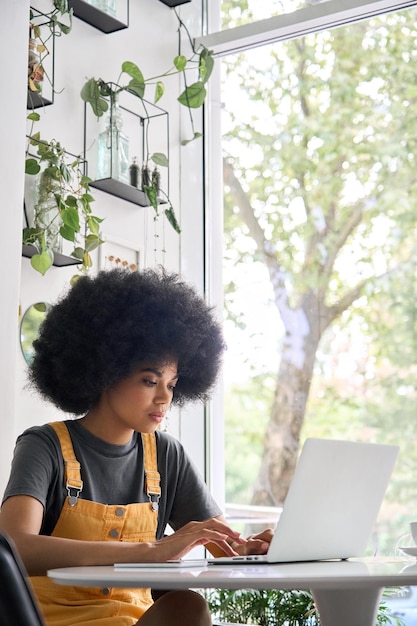 Sérieuse fille noire assise dans un café avec un ordinateur portable en train d'apprendre à travailler en ligne