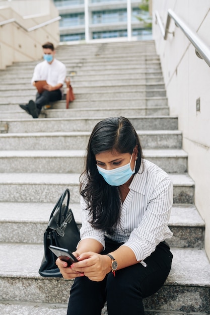 Sérieuse jeune femme d'affaires indienne en masque médical assis sur les marches et la lecture d'un message texte sur l'écran du smartphone