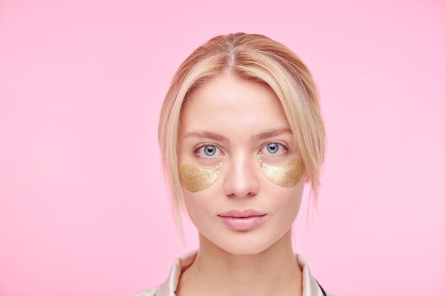 Sérieuse jeune femme blonde avec des patchs sous les yeux revitalisants dorés debout contre le mur rose