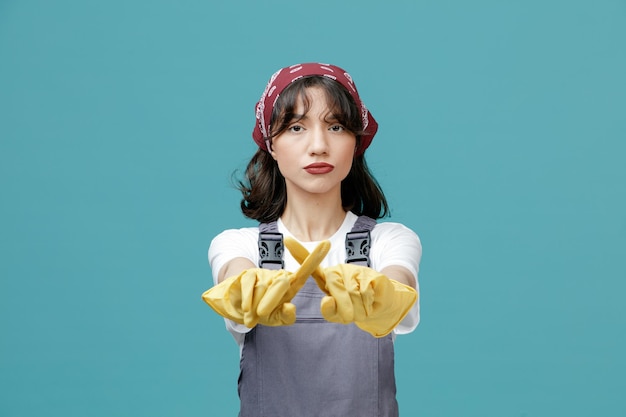 Sérieuse jeune femme nettoyante portant un bandana uniforme et des gants en caoutchouc regardant la caméra s'étirant les mains vers la caméra ne faisant aucun geste isolé sur fond bleu