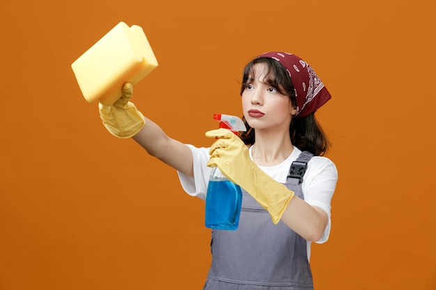 Sérieuse jeune femme nettoyante portant des gants en caoutchouc uniformes et un bandana tenant un nettoyant qui s'étend de l'éponge en regardant le côté faire semblant de nettoyer quelque chose d'isolé sur fond orange