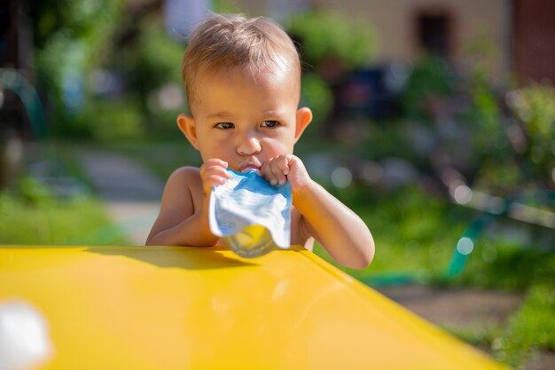 sérieuse petite fille mignonne mangeant de la purée de fruits dans une pochette et regardant la caméra devant la table jaune. sur l'arrière-plan est un jardin verdoyant par une journée ensoleillée en flou