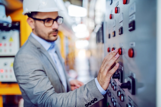 Sérieux beau superviseur caucasien en costume gris et avec casque sur la tête en tournant l'interrupteur. Mise au point sélective à portée de main. Intérieur de la centrale électrique.