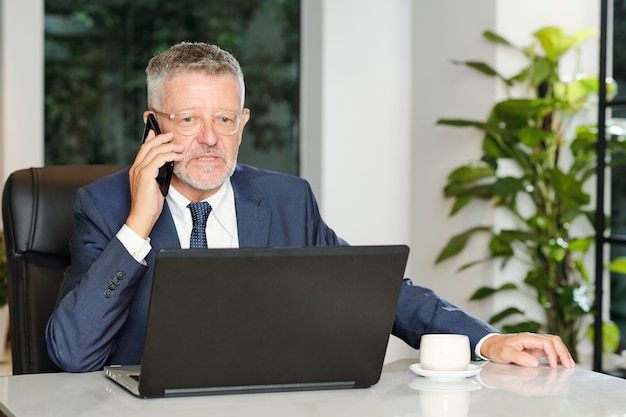 Sérieux entrepreneur senior en colère travaillant sur ordinateur portable et parler au téléphone avec le client