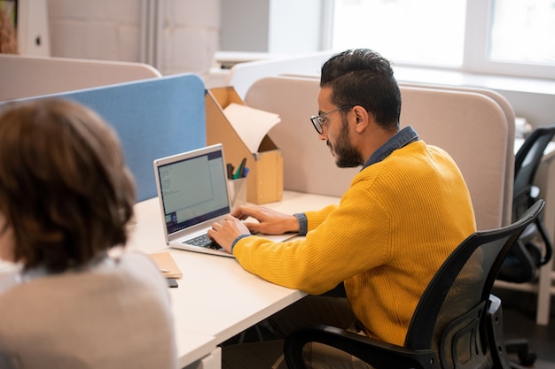 Sérieux Jeune Commerçant Du Moyen-orient Occupé En Pull Jaune Assis Sur Une Chaise Pivotante Et Composer Un Plan De Vente Sur Ordinateur Portable