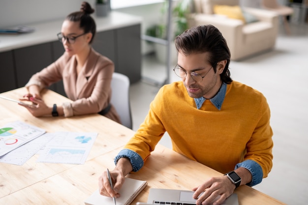 Sérieux jeune gestionnaire en lunettes de prendre des notes tout en analysant les données du site Web sur un ordinateur portable au bureau