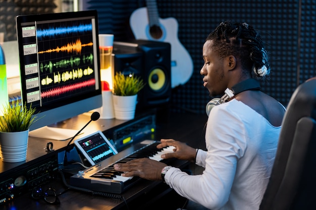 Sérieux Jeune Homme Africain En Appuyant Sur Les Touches Du Clavier De Piano Et En Regardant L'écran De La Tablette Avec Visualisation Sonore De Formes D'onde