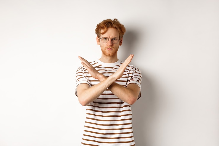 Photo sérieux jeune homme aux cheveux roux fronçant les sourcils, montrant un geste d'arrêt, faisant une croix pour interdire quelque chose de mal, en désaccord avec vous, debout sur fond blanc.