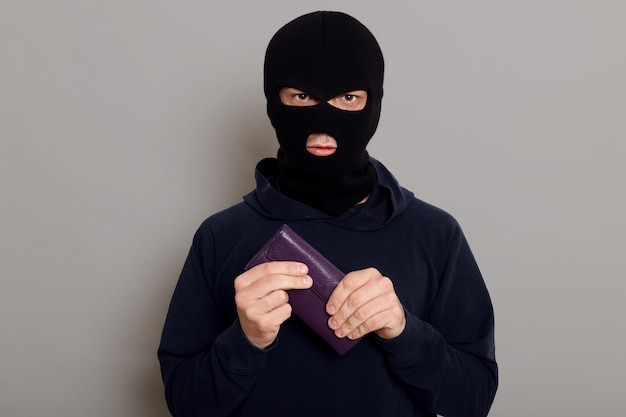 Sérieux jeune homme cambrioleur en masque de vol et pull noir tient fermement le portefeuille qu'il vient de voler