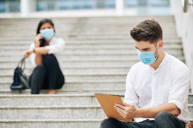 Sérieux jeune homme en masque médical assis sur les marches et lire l'article sur tablette, ses collègues assis en arrière-plan