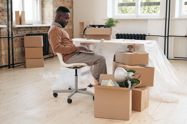 Sérieux jeune homme noir avec barbe assis sur une chaise de bureau et utilisant un ordinateur portable au bureau avec déballé