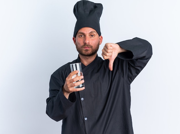 Sérieux jeune homme de race blanche cuisinier en uniforme de chef et casquette tenant un verre d'eau montrant le pouce vers le bas