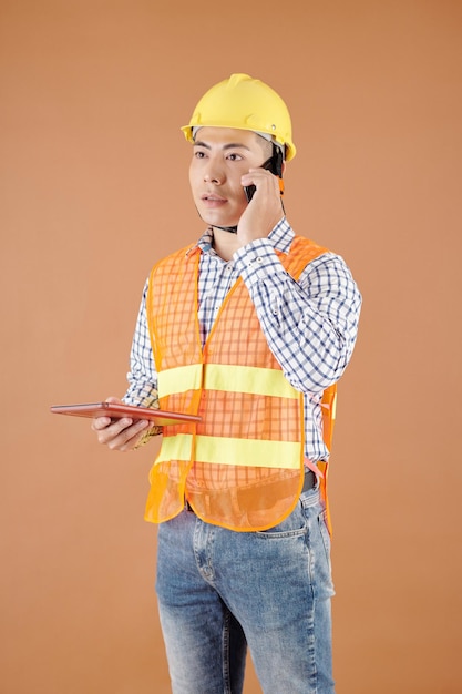 Sérieux jeune ingénieur en construction dans un casque tenant une tablette et parlant au téléphone avec un collègue