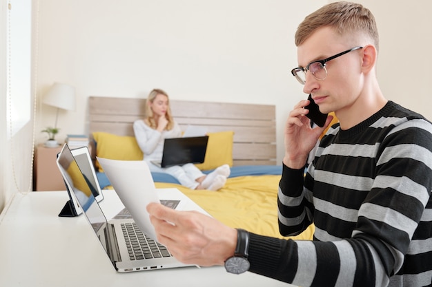 Sérieux jeune ingénieur logiciel discutant d'un document avec un collègue au téléphone lors d'un travail à domicile en raison d'un verrouillage