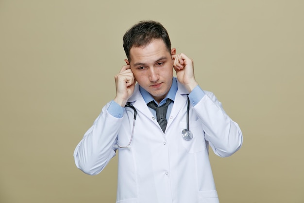 Sérieux jeune médecin de sexe masculin portant une robe médicale et un stéthoscope autour du cou regardant vers le bas tout en mettant les doigts dans les oreilles isolés sur fond vert olive