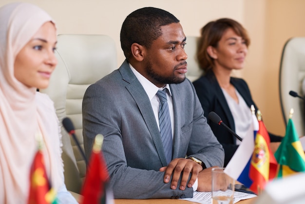 Sérieux jeune orateur afro-américain à l'écoute de l'un de ses collègues étrangers assis entre deux déléguées interculturelles