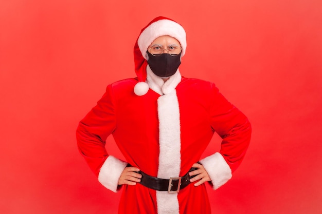 Photo sérieux père noël inquiet dans des lunettes portant un masque facial protecteur regardant la caméra se tenant la main sur les hanches quarantaine pendant les vacances prise de vue en studio intérieure isolée sur fond rouge