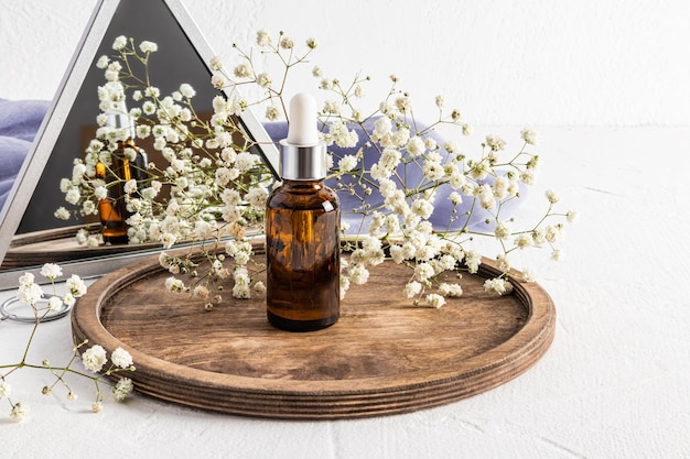 Le sérum de soin de la peau du visage dans une bouteille en verre avec un compte-gouttes se dresse sur un plateau rond en bois avec une gypsophile au miroir des cosmétiques naturels