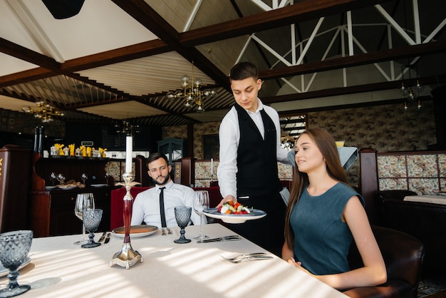 Un serveur élégant sert un jeune couple qui est venu à un rendez-vous dans un restaurant gastronomique. Service à la clientèle dans la restauration.