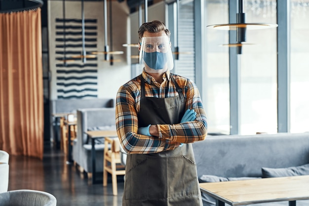 Serveur masculin dans des vêtements de travail protecteurs gardant les bras croisés et tout en se tenant dans le restaurant