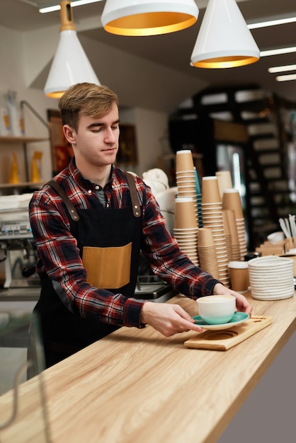 Serveur masculin en tablier tenant une tasse de boisson chaude dans un café, le barista est heureux de travailler à la cafétéria