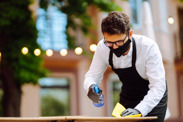 Serveur portant un masque protecteur et des gants nettoyant la table du restaurant pour le prochain client