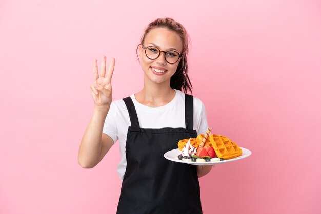 Serveur de restaurant fille russe tenant des gaufres isolées sur fond rose heureux et comptant trois avec les doigts