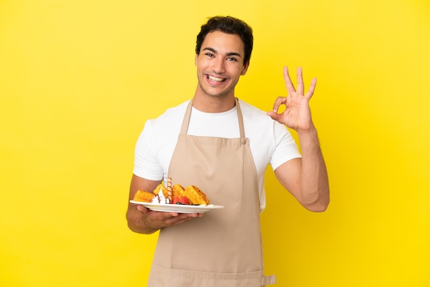 Serveur de restaurant tenant des gaufres sur fond jaune isolé montrant un signe ok avec les doigts