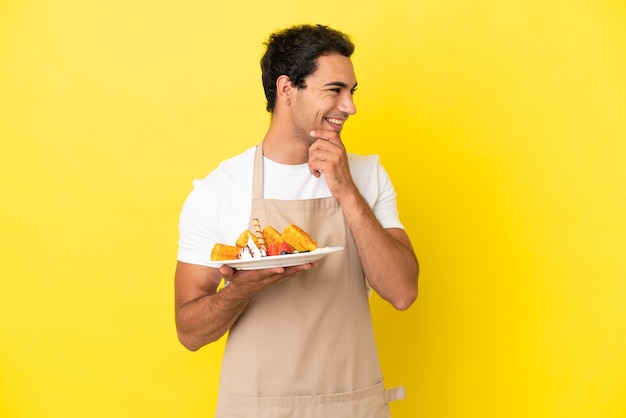 Serveur de restaurant tenant des gaufres sur fond jaune isolé regardant sur le côté et souriant