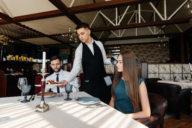 Un serveur stylé sert un jeune couple composé d'un homme et d'une femme venus en rendez-vous dans un restaurant gastronomique Service client dans les établissements de restauration et de restauration