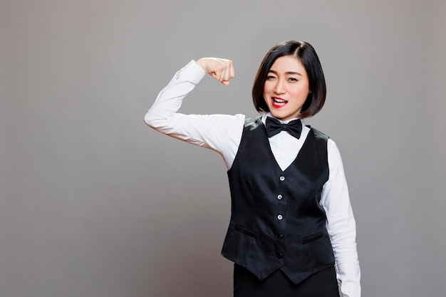 Serveuse asiatique attrayante montrant le concept de force des biceps et regardant la caméra. Jeune réceptionniste forte femme portant un uniforme de restaurant professionnel, portrait musculaire du bras flexible