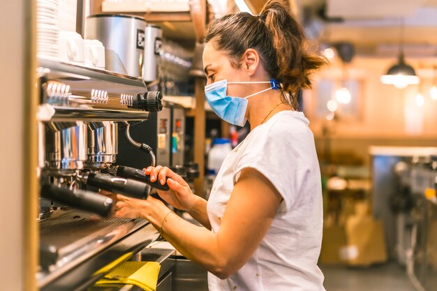 Une serveuse caucasienne aux cheveux noirs avec un masque dans un bar. Préparer un café au lait avec la machine