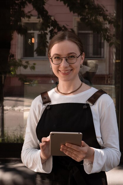 La serveuse se tient à l'extérieur en été avec une tablette dans les mains près de la fenêtre du café