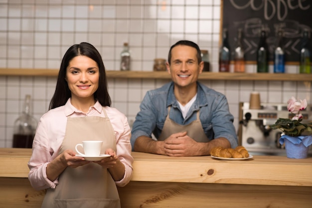 Serveuse souriante tenant une tasse de café et regardant la caméra avec un barista en arrière-plan