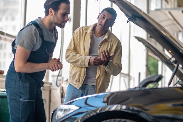 Photo service automobile. afro-américain regardant d'un air interrogateur un mécanicien sérieux caucasien regardant dans un capot de voiture ouvert debout dans un atelier