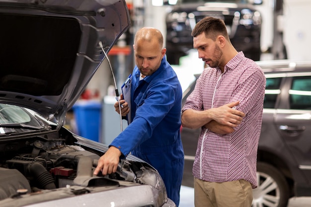 service automobile, réparation, entretien et concept de personnes - mécanicien avec presse-papiers et homme ou propriétaire regardant un moteur de voiture cassé à l'atelier