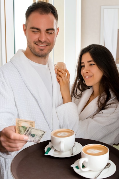 Service de chambre offrant du café dans une chambre d'hôtel pour un couple marié portant un peignoir qui donne des pourboires en dollars