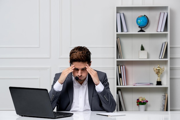 Service client joli gentleman avec ordinateur en costume de bureau gris touchant les tempes très agacé